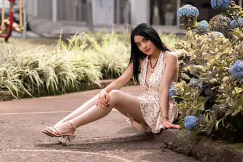 Young woman in a white floral sundress sitting on a garden path near blooming blue hydrangeas.