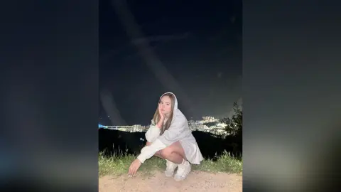 Young woman in a white oversized hoodie sitting on a hill overlooking city lights at night.