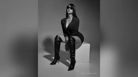 Stylish woman in black blazer and high boots sitting on a white pedestal in a moody black and white studio portrait.