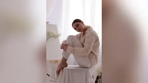 Young woman in beige loungewear sitting on a stool in a bright, minimalist room with a serene mood.