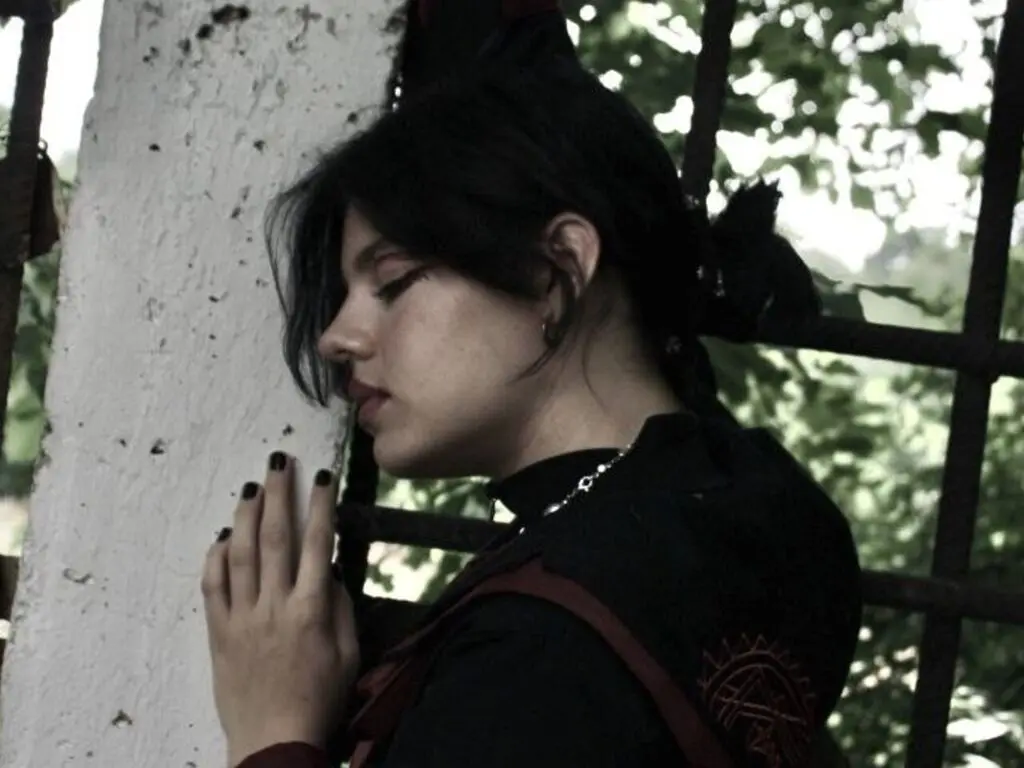 Moody profile portrait of a young woman with dark hair leaning against a white pillar in a leafy garden setting.