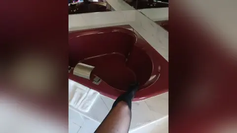 A person in black socks relaxing in a modern red heart-shaped bathtub in a bright marble bathroom.