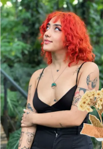 Young woman with curly red hair and tattoos wearing a black camisole in a lush green garden setting.