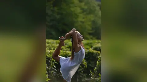 Serene woman in a white summer dress performing a yoga pose in a lush green forest setting.