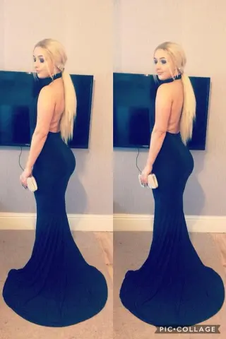 Elegant blonde woman in a backless black evening gown holding a clutch in a modern indoor setting.