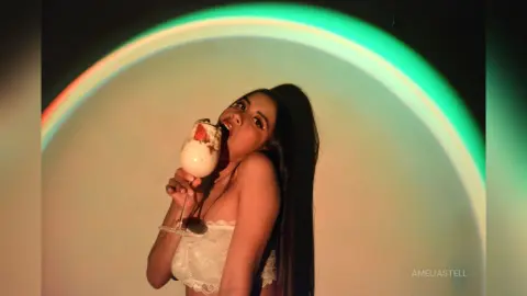 AmeliaStell holds a glass of white cream or foam to her mouth while posing against a bright, circular rainbow-colored light.