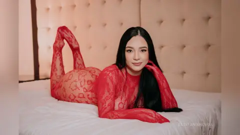 Young woman in a red lace bodysuit lying on a white bed in a cozy bedroom setting.