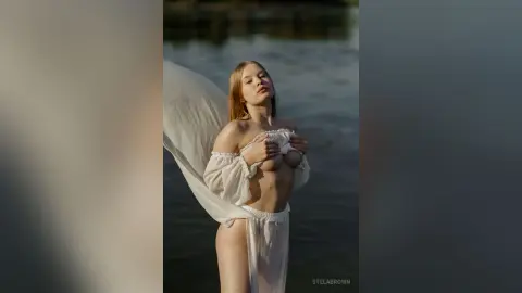StelaBrown poses by the water, holding her white sheer top up to expose her breasts.