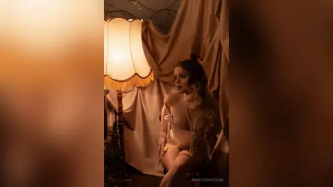 Elegant woman in a satin slip dress posing by a vintage lamp in a warm, moody indoor setting.