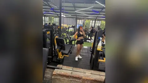 NatiSweet stands in a gym wearing a black sports bra and black shorts, posing next to weight machines.