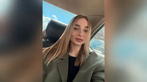 Blonde woman in an olive green coat sitting in a car on a sunny day with a relaxed expression.