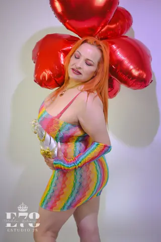 Redheaded woman in a colorful rainbow mini dress posing with large red heart balloons in a bright studio.