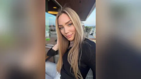 AmiliRay — AmiliRay with long blonde hair smiles while posing in a black long-sleeve top on a balcony.