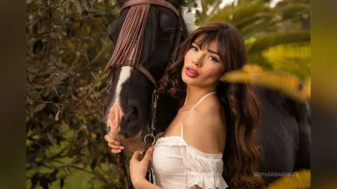 SophiaNapoles poses outdoors with a black horse, wearing a white lace camisole and showing her long brown hair.