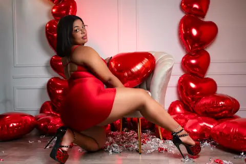 Curvy woman in a red satin dress posing with red heart balloons in a romantic studio setting.