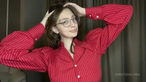 Young woman with glasses wearing a red striped button-down shirt posing in a cozy indoor setting.