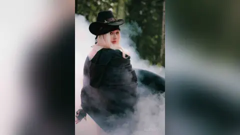 EllieFrosty poses in a black hat and black top or wrap surrounded by thick white smoke or fog.