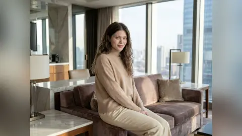 MaryLico sits on a brown velvet sofa wearing a tan turtleneck sweater and light-colored trousers in a bright high-rise apartment.