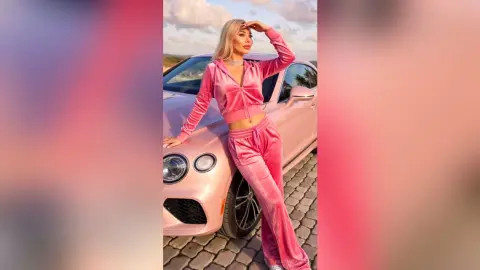 Blonde woman in pink velvet tracksuit leaning on a luxury car during a dreamy sunset.