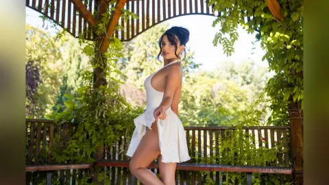 Smiling woman in a white summer dress posing under a lush green garden pergola on a sunny day.