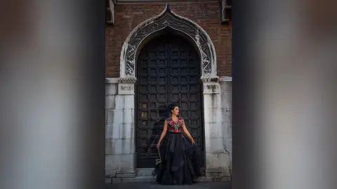 MissyJolie stands before a large, dark arched stone doorway wearing a black sleeveless gown with a red and gold floral bodice.