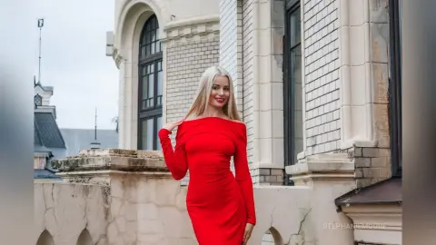 Smiling blonde woman in a long red evening dress standing on a classic stone balcony.