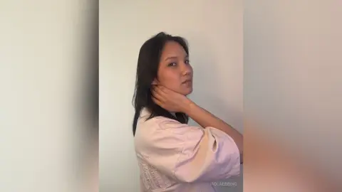 ViolaEbbing poses with her hand on her neck, wearing a light pink shirt against a plain white wall.