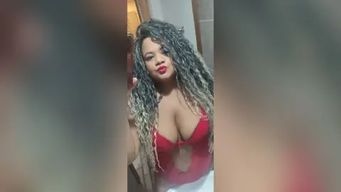 Curvy woman with long wavy blonde hair wearing a red plunging bodysuit in a modern bathroom setting.