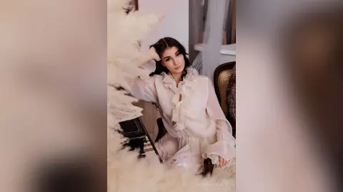 Elegant woman in a white ruffled blouse sitting in a dreamy, soft-lit room with aesthetic decor.
