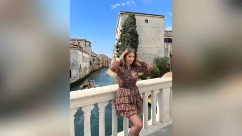 Smiling woman in a floral maxi dress standing on a stone balcony overlooking a scenic Venice canal.