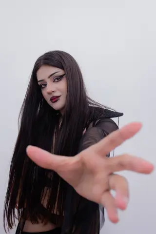 Young woman with long dark hair and bold eyeliner wearing a black sheer outfit against a white background.