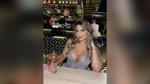 Georgia poses at a bar wearing a low-cut grey top, holding a cocktail with a lime garnish.