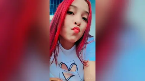 Young woman with vibrant red hair wearing a white graphic t-shirt in a bright, casual indoor setting.