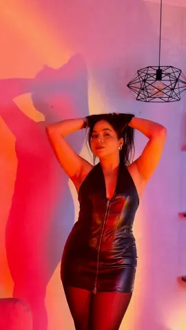 Woman in a black leather halter dress posing against a warm red light background.