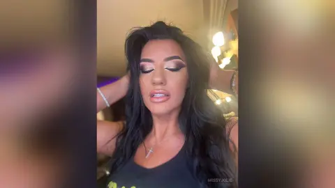 MissyJolie poses with her hands in her long black hair, wearing heavy eye makeup and a black tank top.