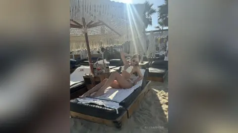 Woman in white bikini relaxing on a beach lounge chair under a boho sunshade on a sunny tropical beach.