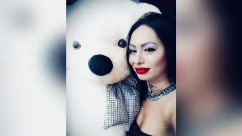 DeeaAnderson poses next to a large white teddy bear wearing a black tank top and bright red lipstick.