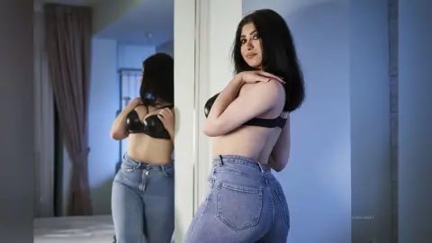Young woman in a black bra and blue jeans posing in a mirror for a moody indoor portrait.