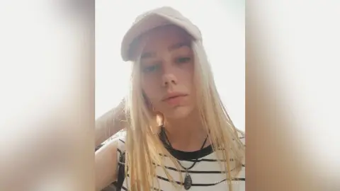 Blonde woman in a beige cap and striped tank top posing for a bright, casual outdoor selfie.