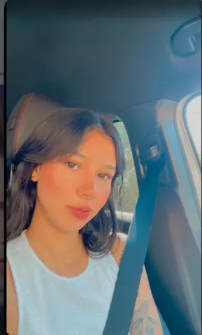 Young woman in a white tank top sitting in a car seat with a relaxed, natural expression.
