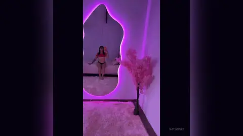 NatiSweet poses in a red crop top and black thong, standing in front of a purple-lit mirror in a room with a pink rug and pink decor.