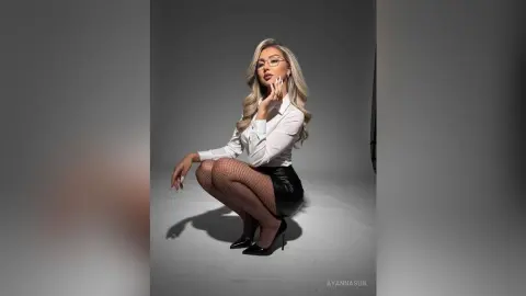Blonde woman in white button-down shirt and black leather skirt posing elegantly in a studio setting.