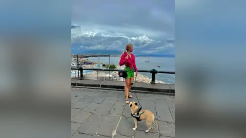 AmaFox — Blonde woman in a pink top and green shorts walking her dog along a scenic coastal promenade under cloudy skies.