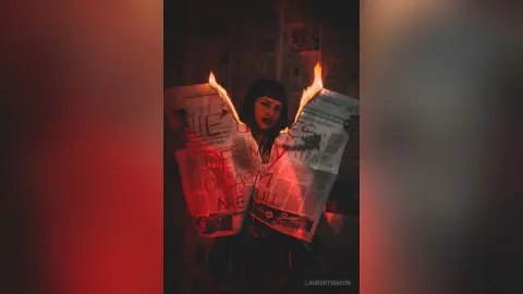 Moody portrait of a woman in dark clothing holding burning newspapers with red dramatic lighting.