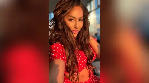 Kayla poses with long wavy brown hair, wearing a red and white polka dot top or dress.