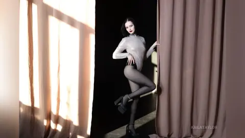 Elegant woman in a silver bodysuit and black tights posing by curtains in dramatic studio lighting.