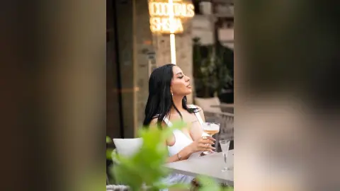 NicoleBella poses outdoors with long black hair, wearing a white tank top and holding a cocktail glass.
