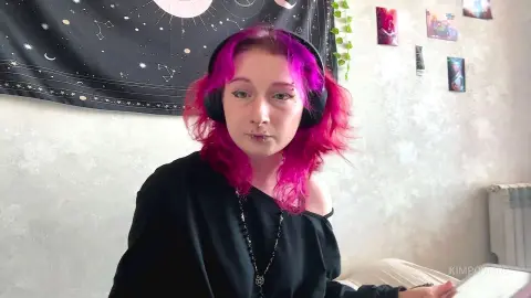 Young woman with pink hair and headphones wearing a black top in a cozy bedroom setting.