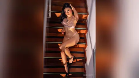 RegineJeans poses on a wooden staircase wearing a sheer pink lace lingerie set and black high heels.