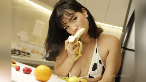Young woman in a white polka dot bikini eating a banana in a bright kitchen setting.